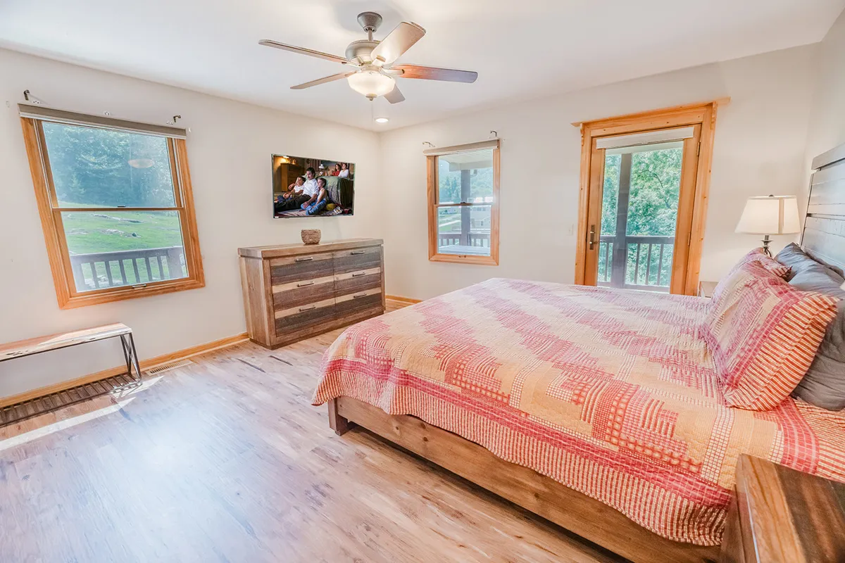 A bright, inviting bedroom with a comfortable bed topped with plush linens, a dresser, large windows letting in natural light, and a wall-mounted television for entertainment. The serene setting makes it ideal for relaxation and unwinding after a long day.