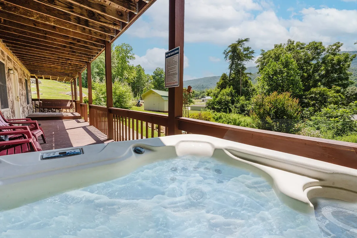 A spacious deck featuring a hot tub positioned to take full advantage of lush greenery and distant mountain views. The scene is tranquil, inviting guests to relax in the bubbling water while enjoying the scenic backdrop. Ample deck space and the presence of outdoor seating suggest it’s perfect for both relaxing mornings and social evenings.
