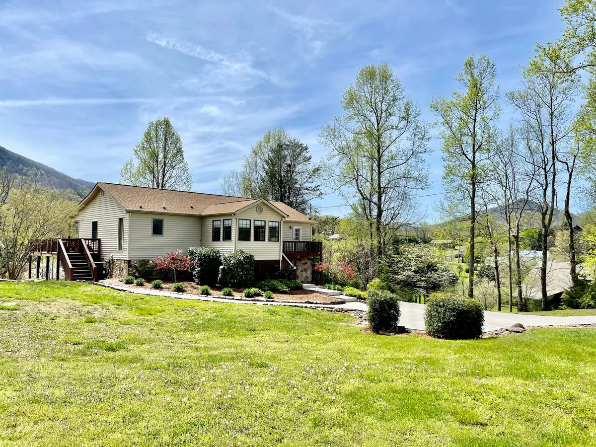 A wide view of the house, showcasing the deck, lush green lawn, and surrounding trees. The spacious lawn and deck provide visual cues for outdoor relaxation, family gatherings, and a sense of being close to nature—key aspects when exploring or returning from local attractions.