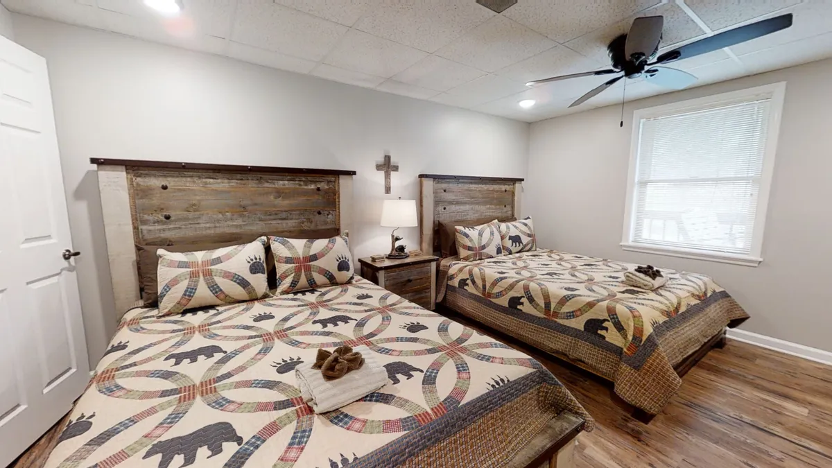 A spacious lower-level bedroom featuring two queen-size beds with rustic wooden headboards and colorful wildlife-themed quilted bedspreads. A nightstand with a lamp and decorative cross adds a homey touch, and sunlight filters through the window to create a welcoming space.
