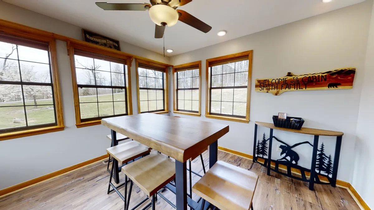 A large dining table surrounded by wooden chairs, with a 'Welcome to the Cabin' sign and expansive windows offering views and natural light. The table is set for family meals, game nights, and group celebrations.
