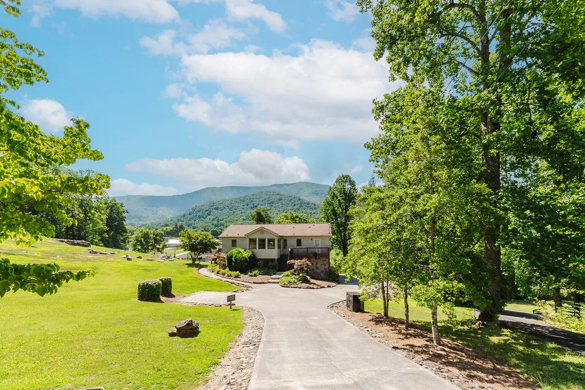 The image captures the exterior of Cove Mountain Rest, with the house nestled among trees and set against a breathtaking mountain backdrop. The lush green lawn and expansive driveway set the stage for outdoor activities and gatherings, visually communicating a sense of arrival and adventure.