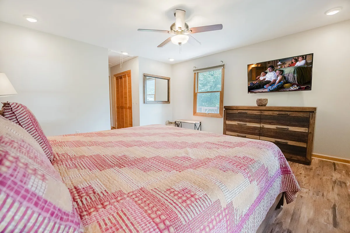 A cozy bedroom scene with a bed dressed in a patterned quilt, a wooden dresser, a wall-mounted television, and a large window flooding the room with natural light. The ambiance is quiet and restful, ideal for unwinding with a good book or movie.