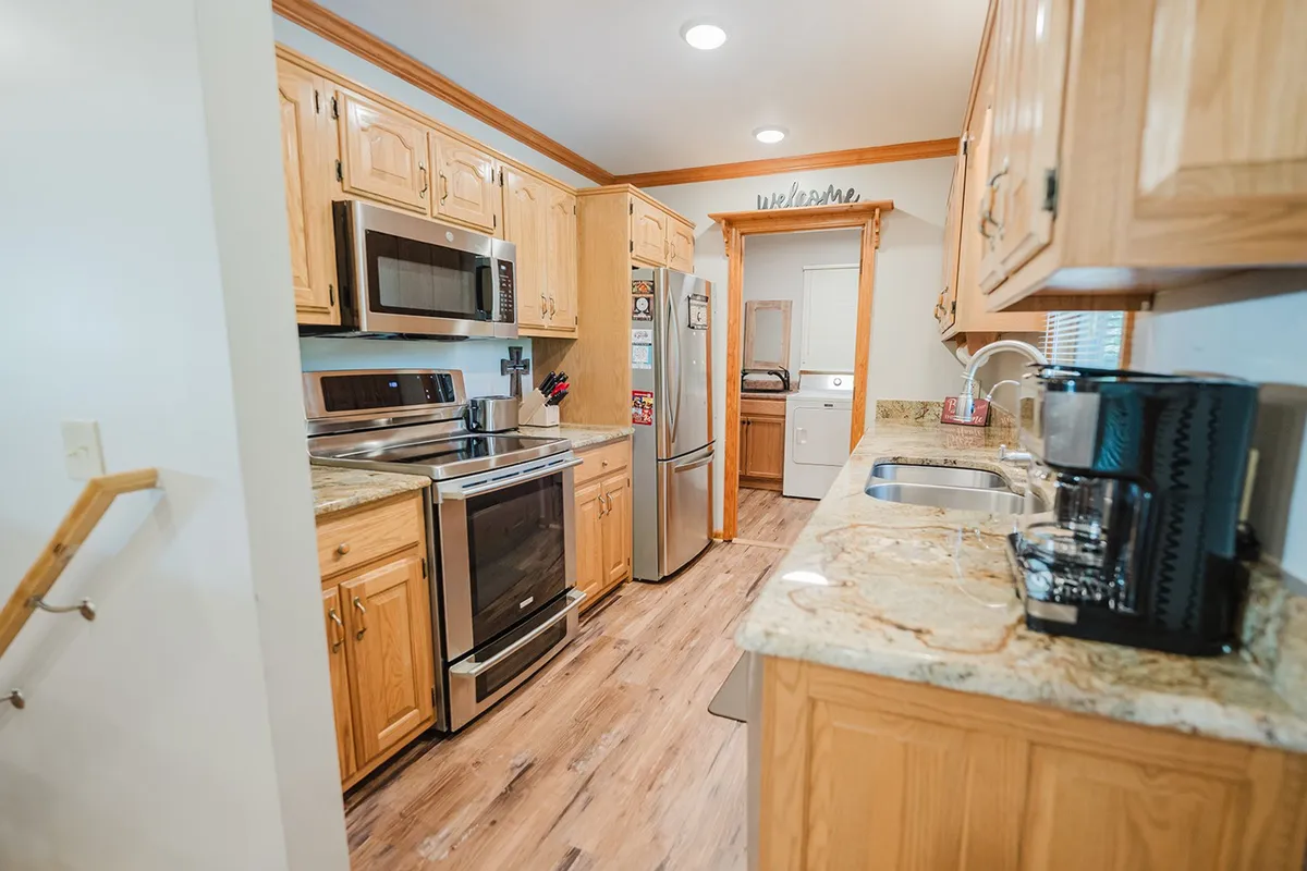 A warmly lit kitchen featuring stainless steel appliances, granite countertops, wooden cabinetry, and a coffee maker. The scene evokes a spacious, well-equipped environment ideal for preparing group meals and gathering for breakfast or dinner.