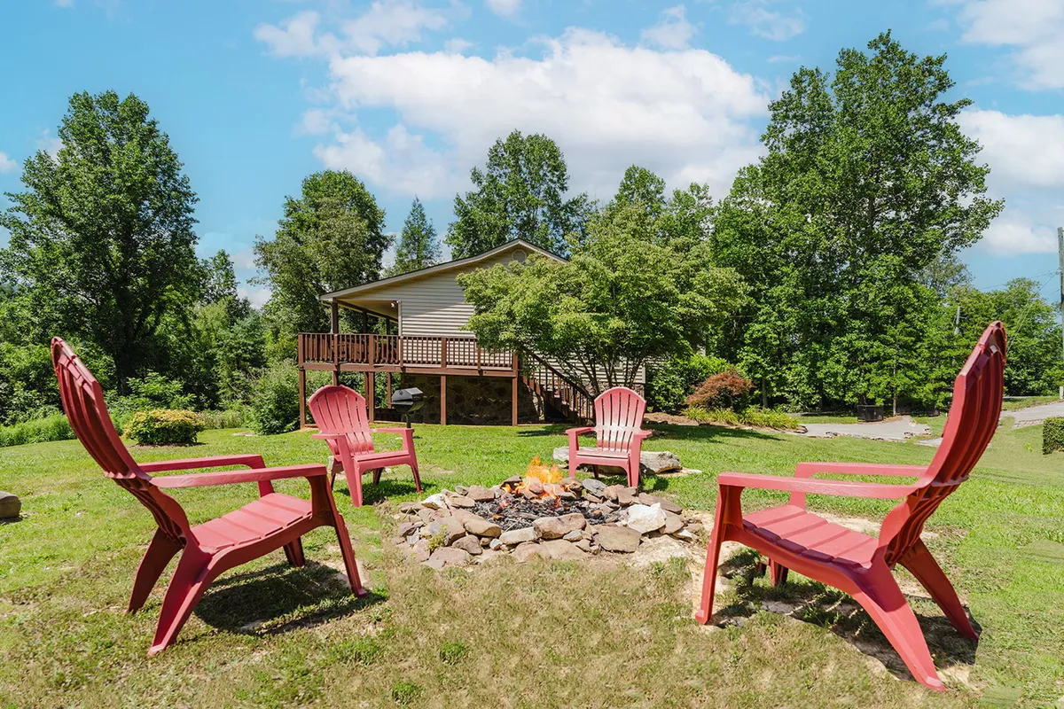 A cozy fire pit surrounded by red chairs sits in the foreground, with the house visible behind. The scene suggests evenings spent making s'mores, sharing stories, and relaxing together outdoors under the stars.