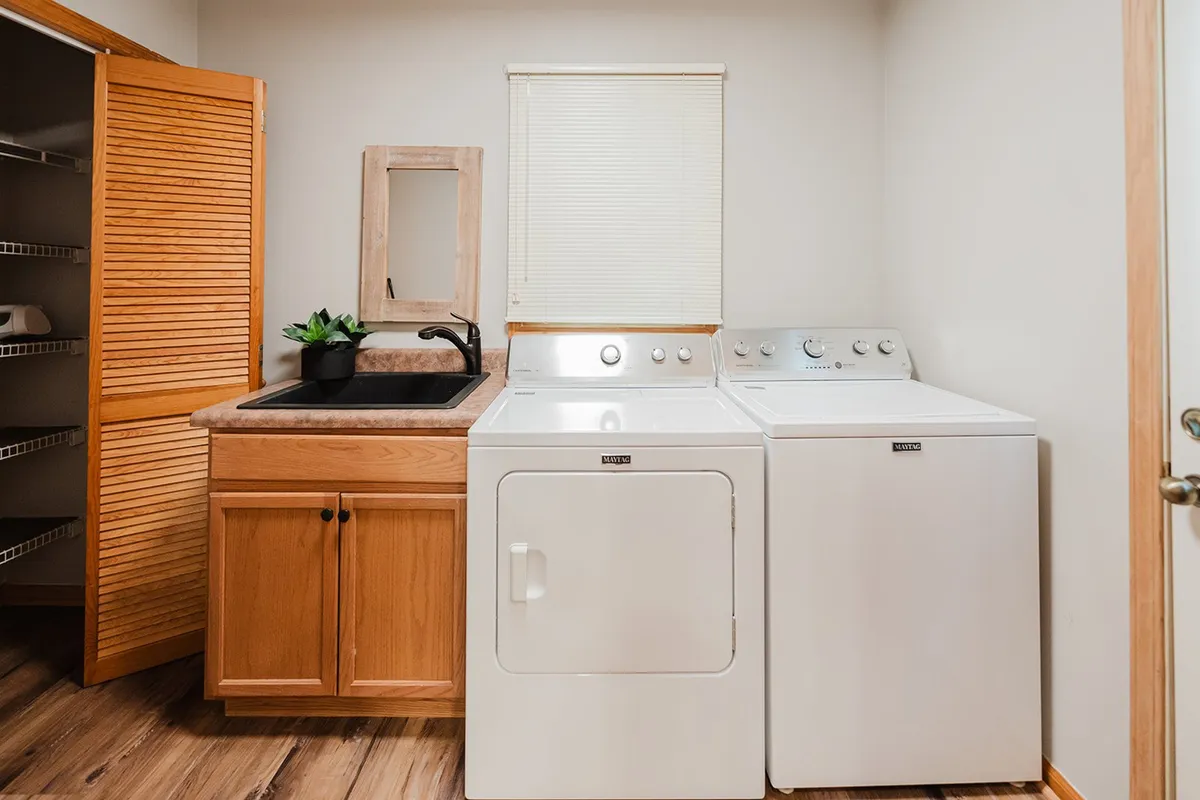 A practical laundry/mudroom featuring a washing machine, dryer, utility sink, wooden cabinets, and a cheerful potted plant. The space feels efficient yet inviting, perfect for managing chores during extended group stays.
