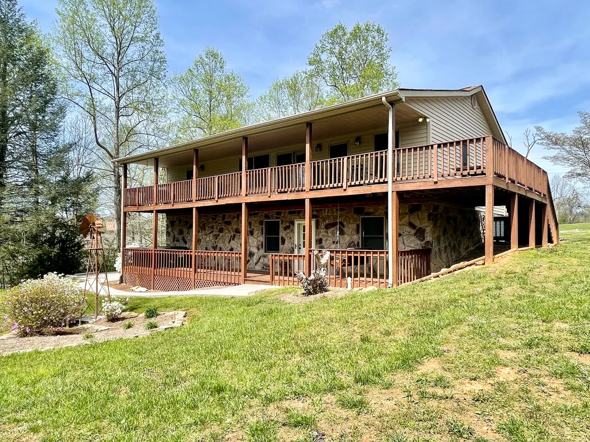 A wide wooden deck extends toward a lush green lawn bordered by trees, providing plenty of space for children to play or families to gather. The deck’s stone facade adds rustic charm, while the overall setting is peaceful and welcoming.