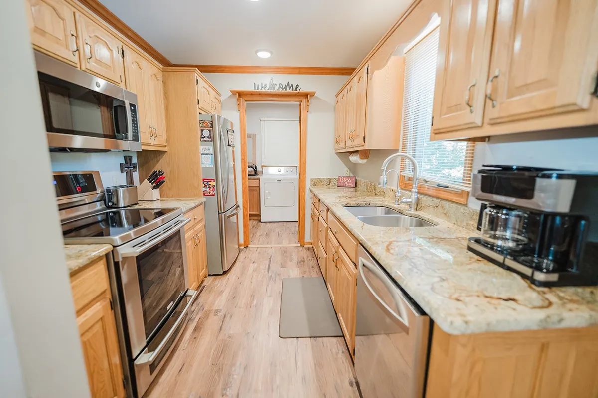 A view of a kitchen with granite countertops, stainless steel appliances, wood cabinets, and large windows allowing natural light to flood the space. The inviting setting is perfect for cooking, baking, and hosting family gatherings.