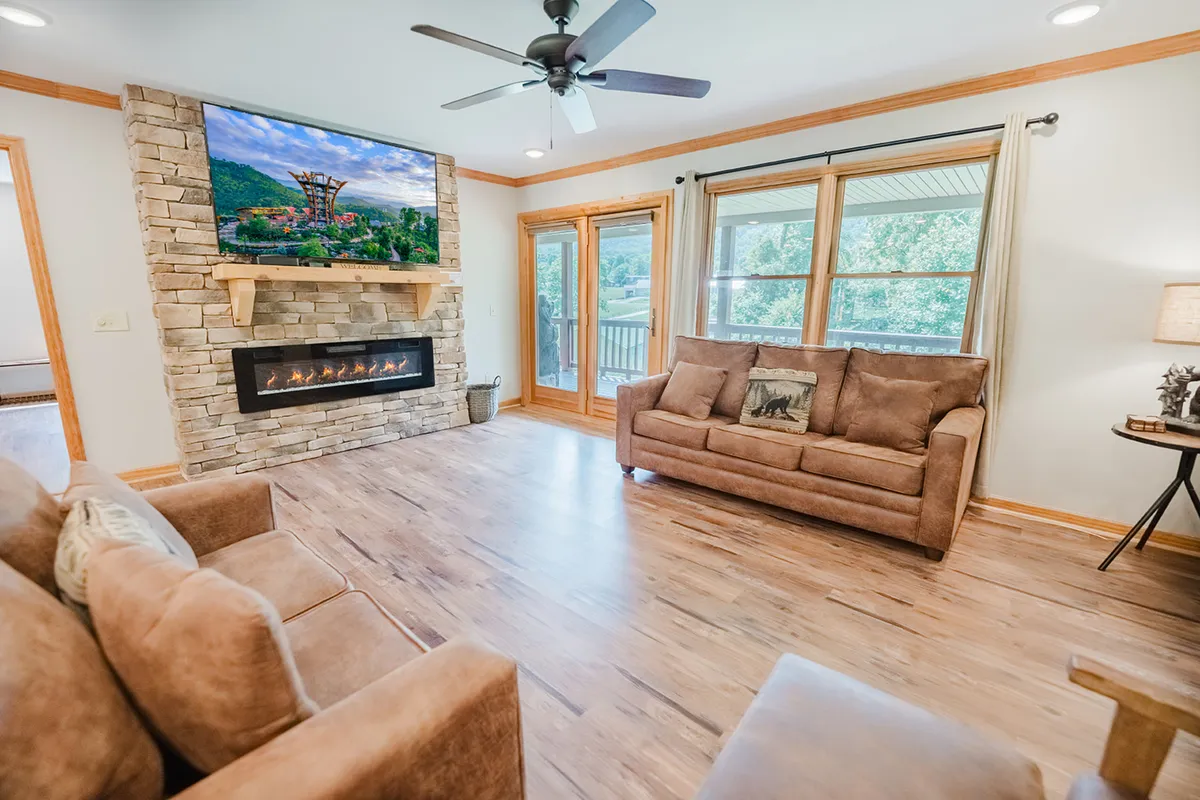 A spacious, sunlit living room featuring a striking stone fireplace, a large television mounted above it, plush and comfortable seating, and expansive windows that let in natural light and offer views of the outdoors. The arrangement invites social gatherings, relaxation, and cozy evenings.